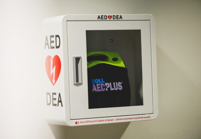 Un défibrillateur externe automatisé (DEA) monté au mur, dans une armoire blanche avec une porte transparente, montrant un appareil 'Zoll AED Plus' à l'intérieur