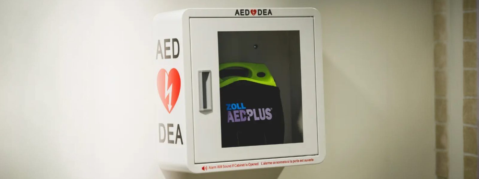 Un défibrillateur externe automatisé (DEA) monté au mur, dans une armoire blanche avec une porte transparente, montrant un appareil 'Zoll AED Plus' à l'intérieur